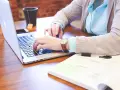 Kobieta pracuje na laptopie, szukając informacji, jak wyrobić paszport online. Na biurku leży notatnik i kubek z herbatą.