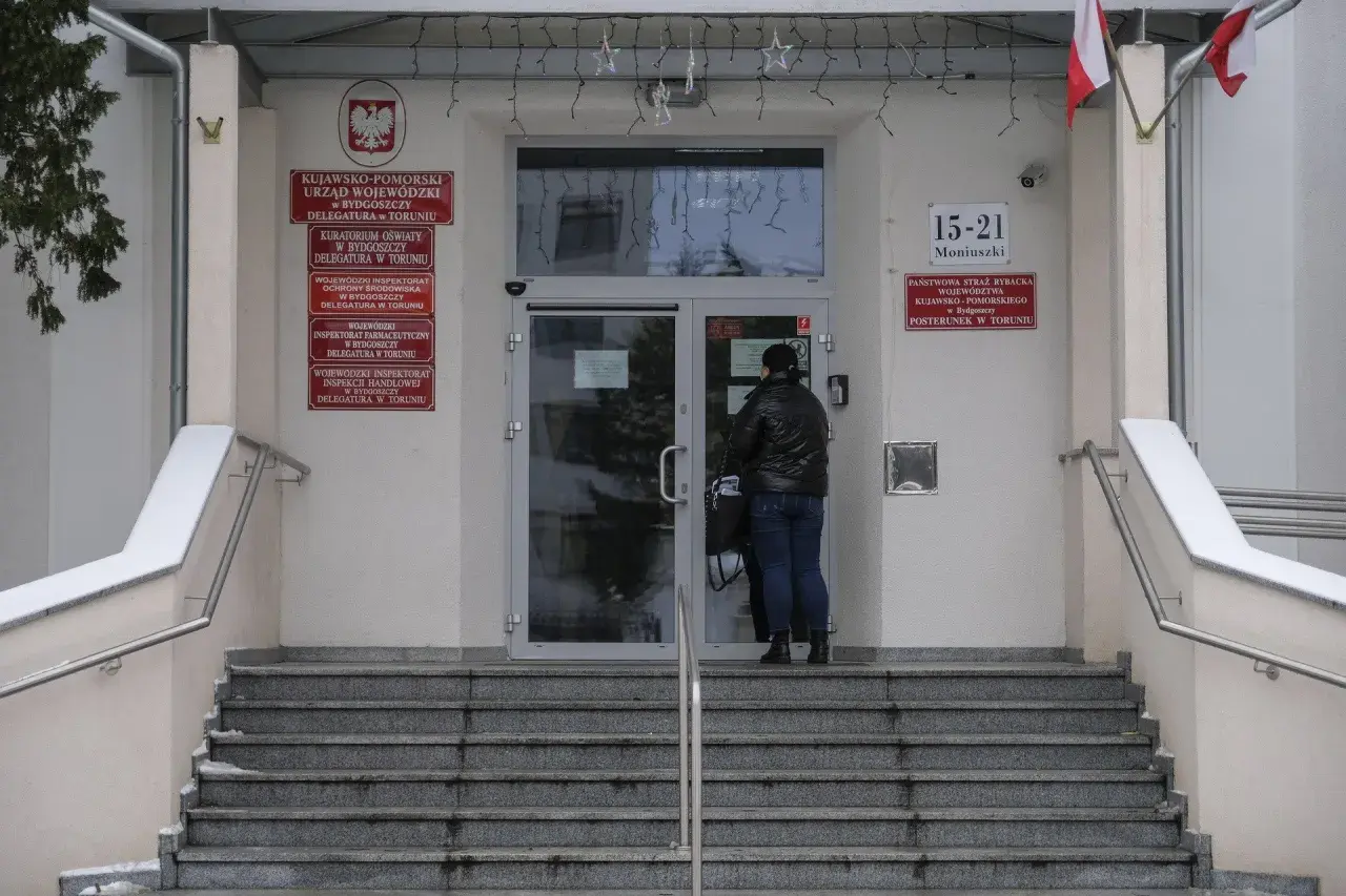 Kobieta przed wejściem do urzędu paszportowego Toruń. Na budynku tablice informacyjne, m.in. Urząd Wojewódzki.
