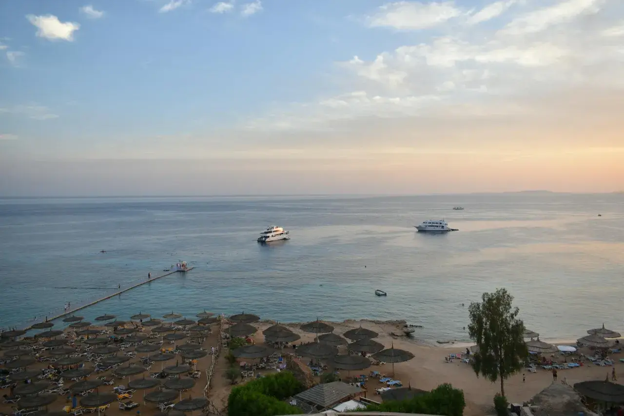 Spokojna plaża z leżakami i parasolami, statki na morzu. Czy wyjazd do Sharm El Sheikh jest bezpieczny? Widok sugeruje relaks.