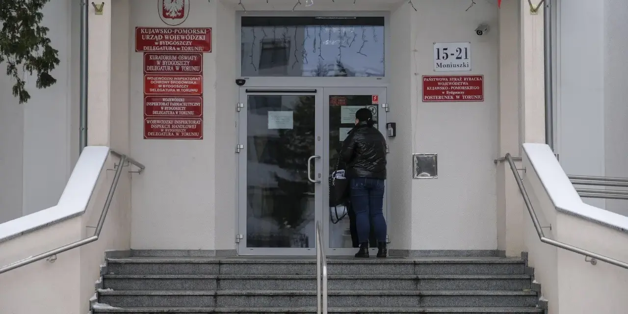 Kobieta przed wejściem do urzędu paszportowego Toruń. Na budynku tablice informacyjne, m.in. Urząd Wojewódzki.