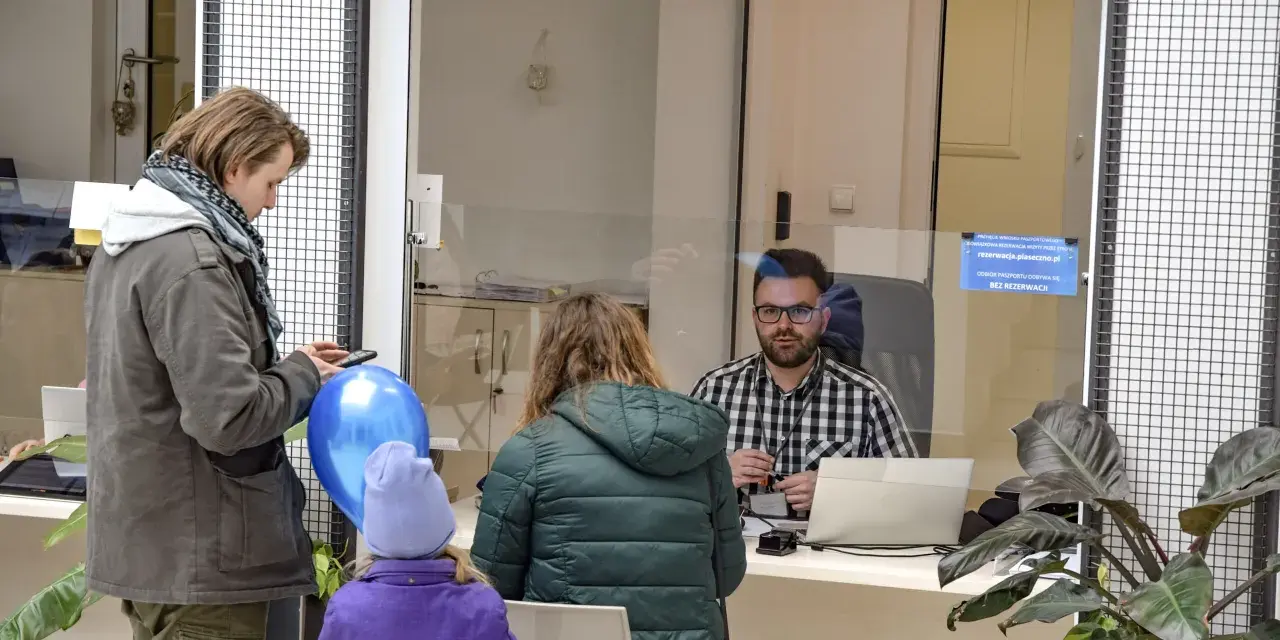 Rodzina czeka na obsługę w urzędzie paszportowym Piaseczno. Mężczyzna z telefonem i niebieskim balonem, kobieta z dzieckiem w fioletowej kurtce.