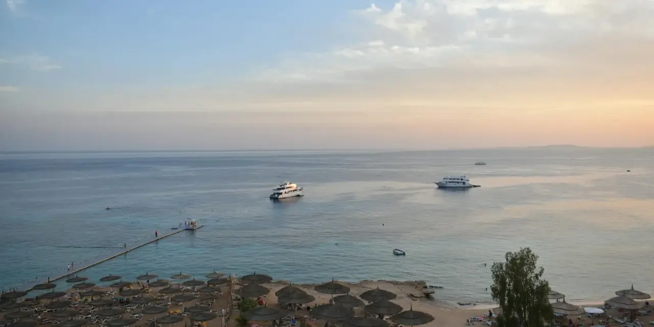 Spokojna plaża z leżakami i parasolami, statki na morzu. Czy wyjazd do Sharm El Sheikh jest bezpieczny? Widok sugeruje relaks.