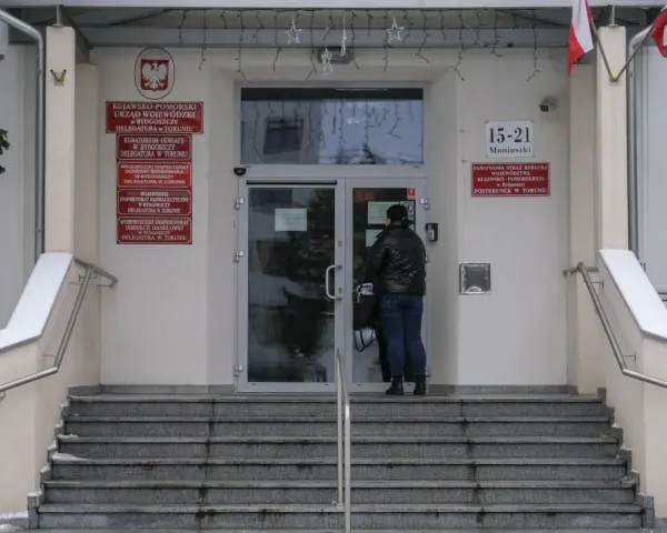 Kobieta przed wejściem do urzędu paszportowego Toruń. Na budynku tablice informacyjne, m.in. Urząd Wojewódzki.