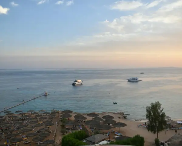 Spokojna plaża z leżakami i parasolami, statki na morzu. Czy wyjazd do Sharm El Sheikh jest bezpieczny? Widok sugeruje relaks.