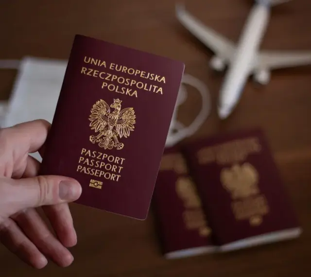 Dłoń trzyma paszport RP. W tle maseczka, samolot i kolejne paszporty, jakby przygotowania do podróży z urzędu wojewódzkiego w Kielcach.