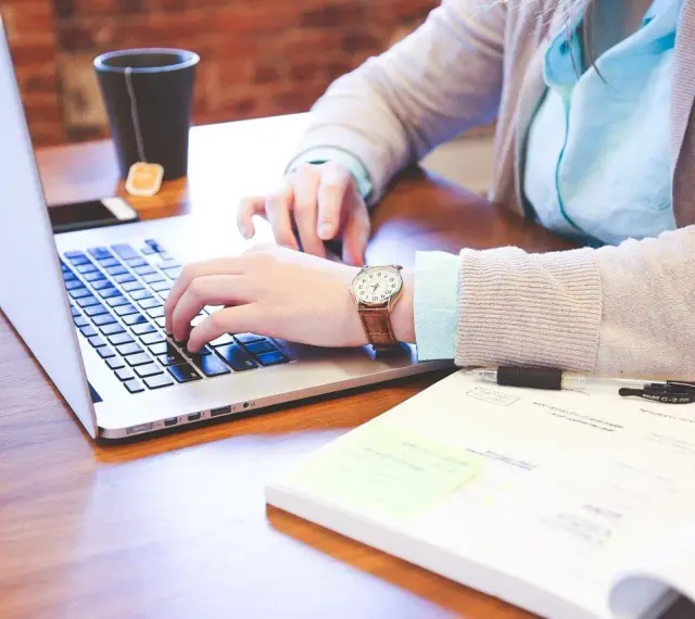 Kobieta pracuje na laptopie, szukając informacji, jak wyrobić paszport online. Na biurku leży notatnik i kubek z herbatą.