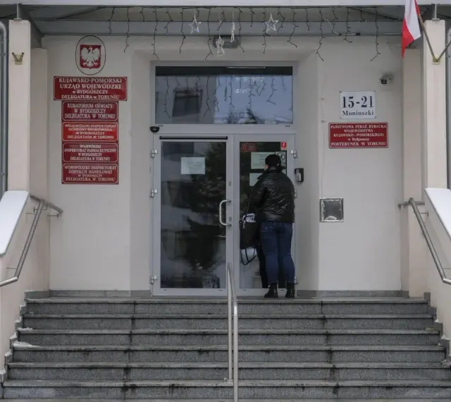 Kobieta przed wejściem do urzędu paszportowego Toruń. Na budynku tablice informacyjne, m.in. Urząd Wojewódzki.