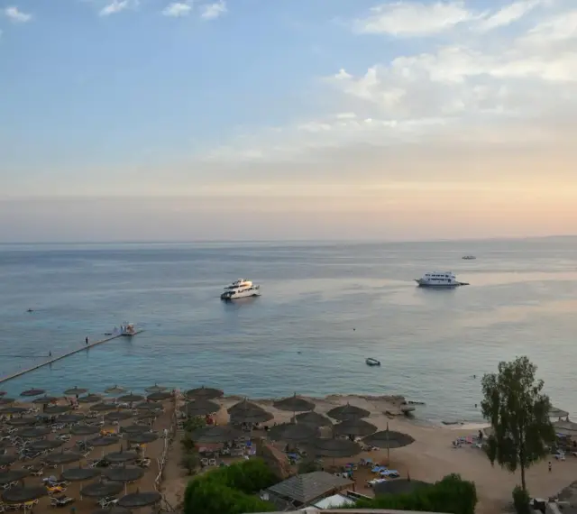 Spokojna plaża z leżakami i parasolami, statki na morzu. Czy wyjazd do Sharm El Sheikh jest bezpieczny? Widok sugeruje relaks.