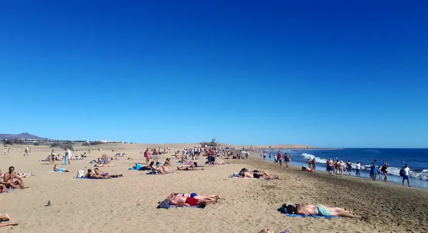 Tłum ludzi odpoczywa na słonecznej plaży. Woda jest spokojna, a niebo bezchmurne. Czy Gran Canaria jest bezpieczna? Tak, wygląda na to, że turyści czują się tu swobodnie.