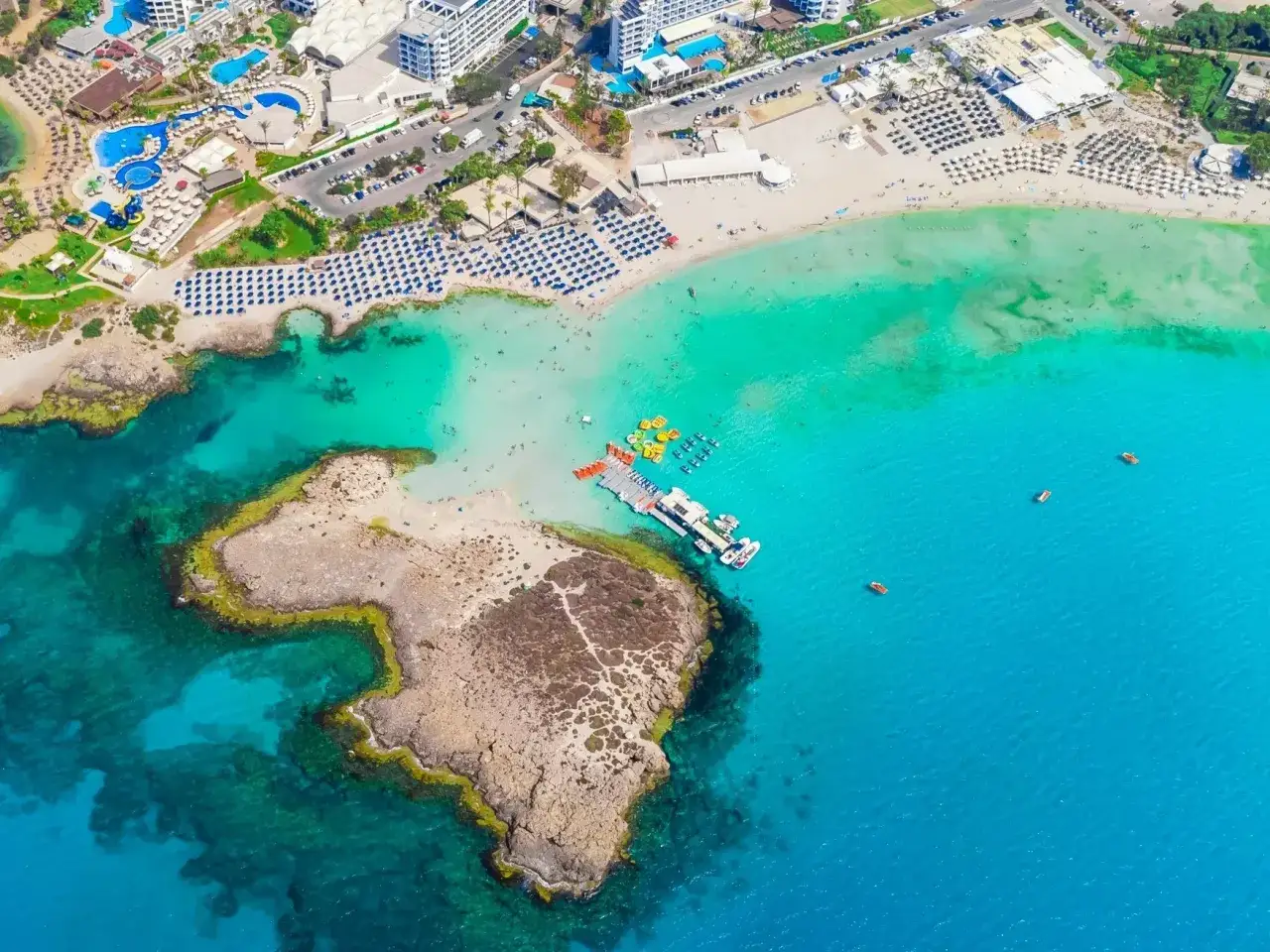 Malownicza plaża na Cyprze z turkusową wodą, hotelami i leżakami. Widać łodzie i skaliste wybrzeże, co świadczy o bezpieczeństwie na Cyprze dla turystów.