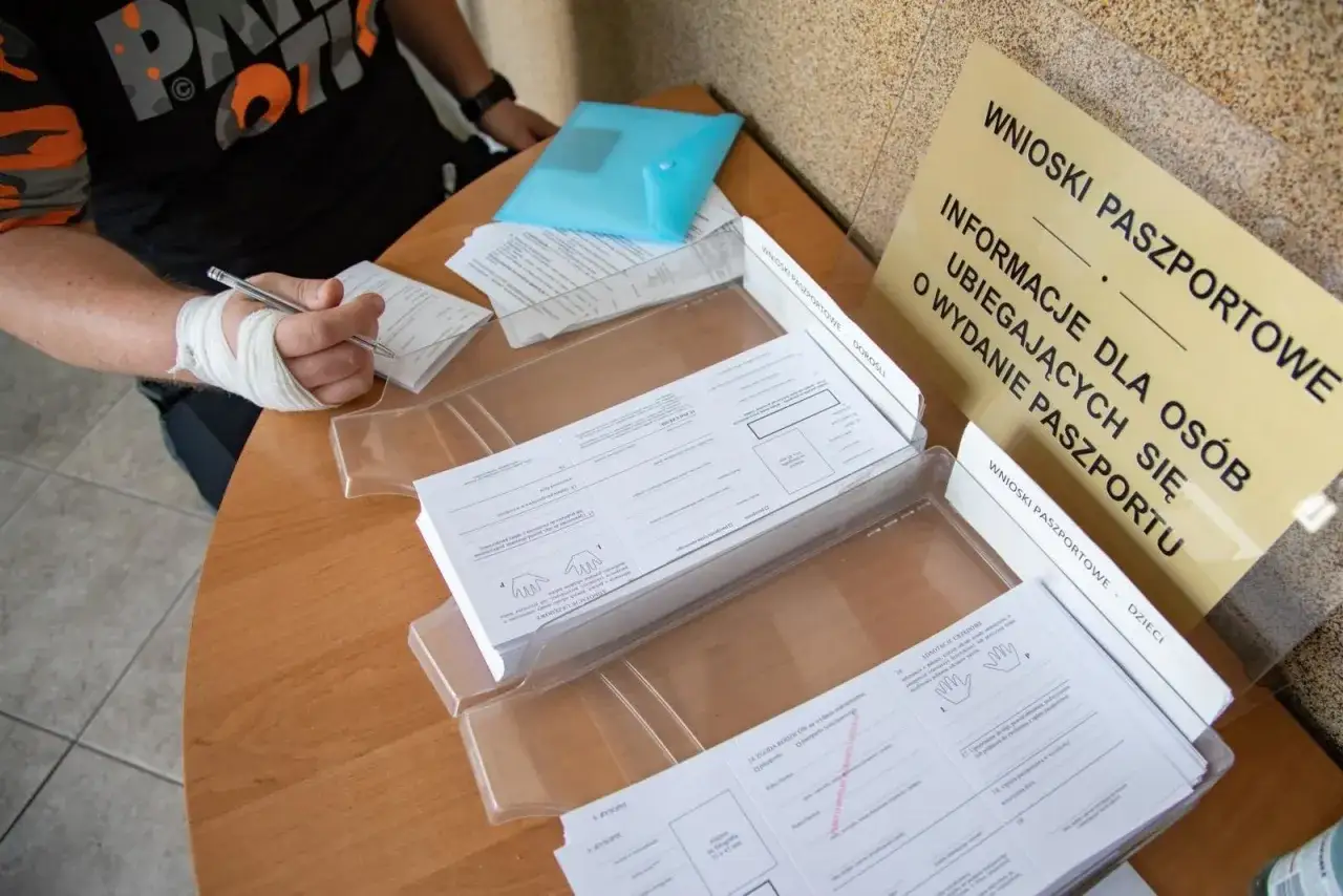 Osoba z zabandażowaną ręką wypełnia wniosek o paszport w Gdyni. Na stole formularze i tablica informacyjna.