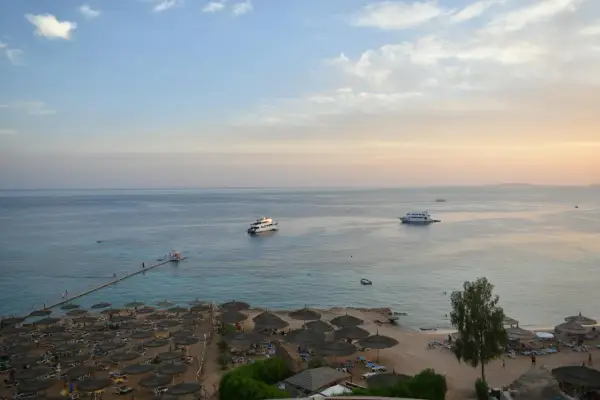 Spokojna plaża z leżakami i parasolami, statki na morzu. Czy wyjazd do Sharm El Sheikh jest bezpieczny? Widok sugeruje relaks.