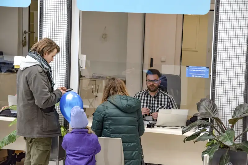 Rodzina czeka na obsługę w urzędzie paszportowym Piaseczno. Mężczyzna z telefonem i niebieskim balonem, kobieta z dzieckiem w fioletowej kurtce.