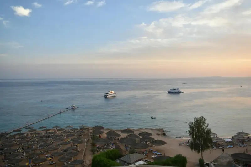 Spokojna plaża z leżakami i parasolami, statki na morzu. Czy wyjazd do Sharm El Sheikh jest bezpieczny? Widok sugeruje relaks.